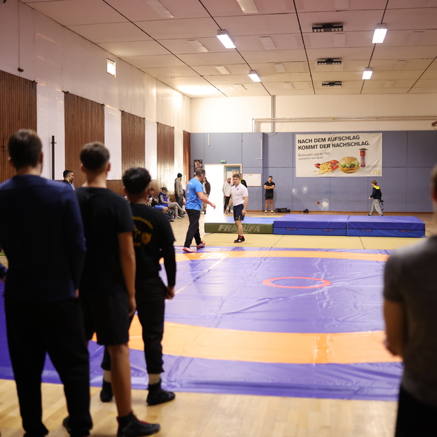 Trainingssession in einem Sportraum mit mehreren Personen, die auf einer blauen Mattenfl&auml;che Wrestling-Techniken &uuml;ben.