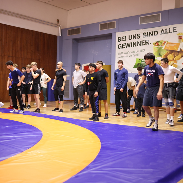 Trainingsgruppe von jungen M&auml;nnern auf einer Ringsportmatte in einer Sporthalle, Hintergrund mit Werbung an der Wand.