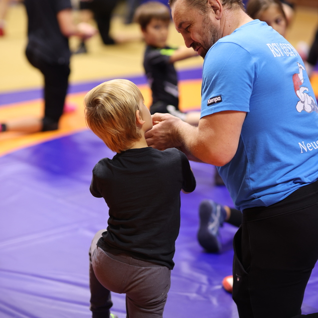 Trainer erkl&auml;rt einem Kind auf einer Tatami-Platte Grundtechniken des Kampfsports. Weitere Kinder &uuml;ben im Hintergrund.