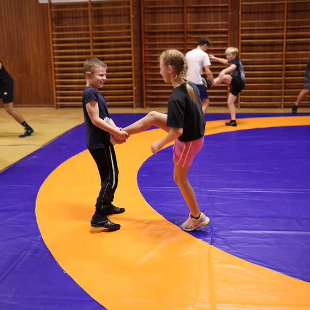 Zwei Kinder &uuml;ben im Trainingsraum miteinander, w&auml;hrend im Hintergrund weitere Personen am Mattenboden trainieren.
