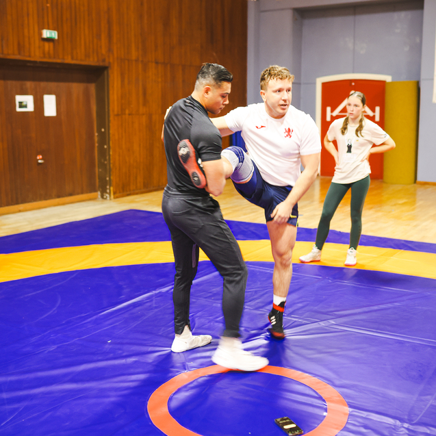 Zwei Personen &uuml;ben einen Wrestling-Move auf einer blauen Matte, w&auml;hrend andere Zuschauer zuschauen.