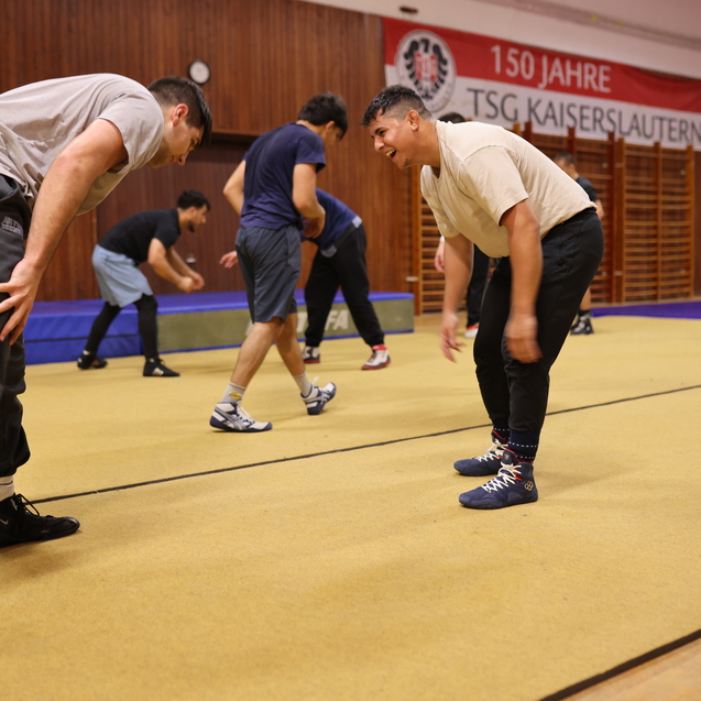 Zwei Sportler in Trainingskleidung beugen sich in einer Halle, w&auml;hrend andere im Hintergrund Bewegungstraining ausf&uuml;hren.