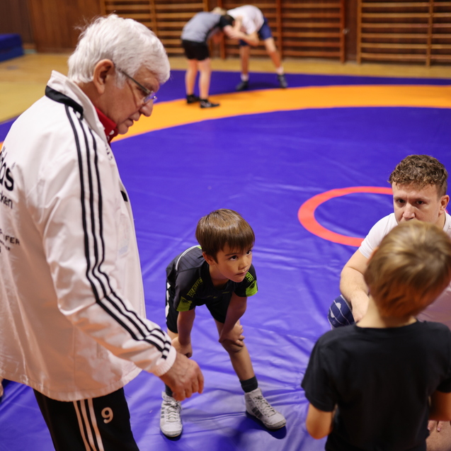 Eine Gruppe von Kindern und Trainern bespricht Technik auf einer blauen Ringermatte in einem Sportraum.