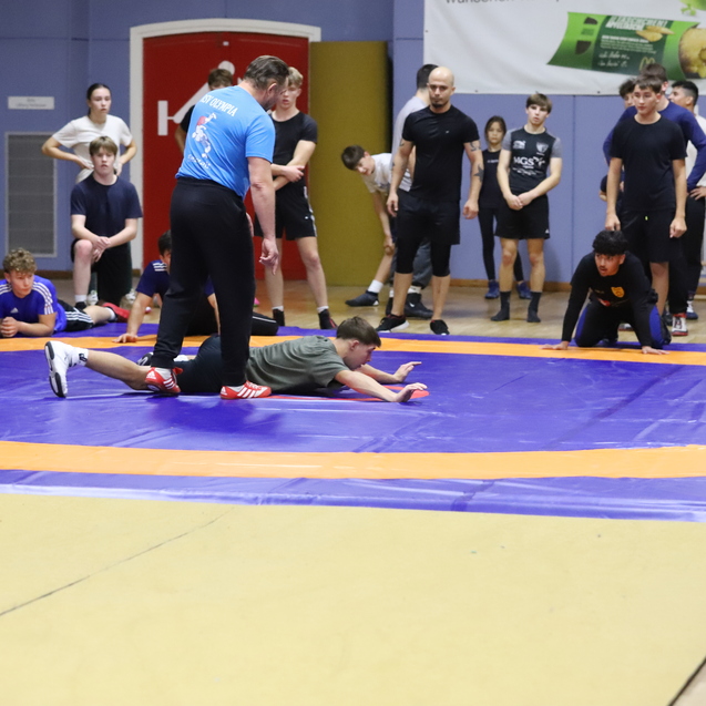 Zweikampftraining in einer Sporthalle, mit einem Trainer und einem K&auml;mpfer auf einer lila Ringfl&auml;che, Zuschauer im Hintergrund.