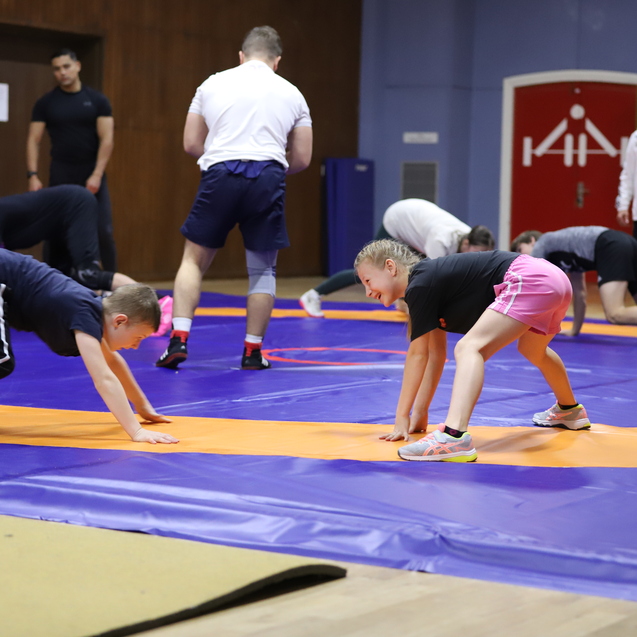 Kinder und Jugendliche &uuml;ben in einer Sporthalle auf einer lila-orangefarbenen Matte im Ringen.
