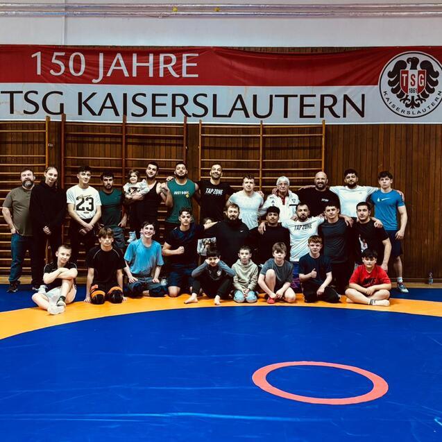 Gruppenfoto von Wrestlern auf einer blauen Matte, im Hintergrund ein Banner der TSG Kaiserslautern zum 150-j&auml;hrigen Jubil&auml;um.