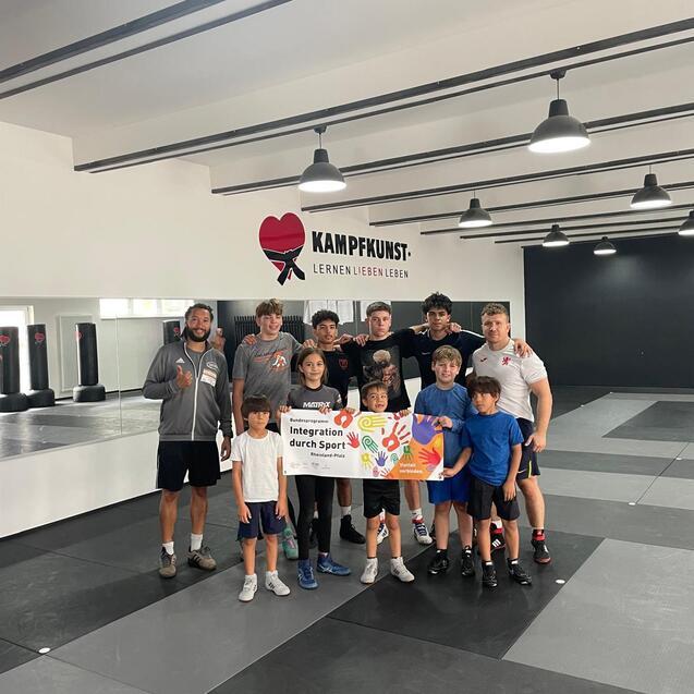 Gruppe junger Sportler mit Trainern h&auml;lt ein Banner zur Integration durch Sport in einem Trainingsraum.