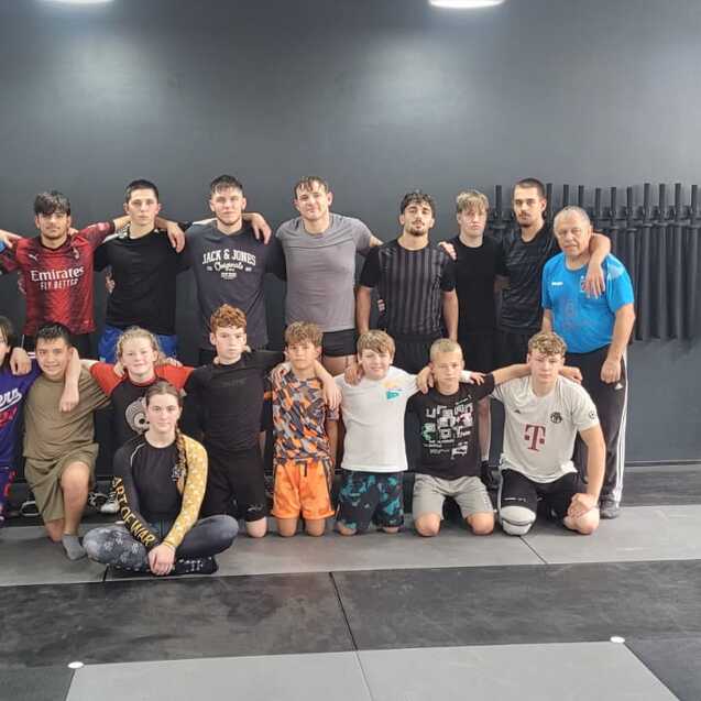 Gruppe von 17 Personen nach dem Training in einem Sportstudio, umgeben von Matten und Trainingsger&auml;ten.