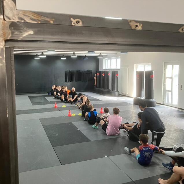 Blick durch die T&uuml;r auf eine Gruppe von Sportlern, die auf Matten in einem Trainingsraum sitzen, w&auml;hrend ein Trainer anleitet.