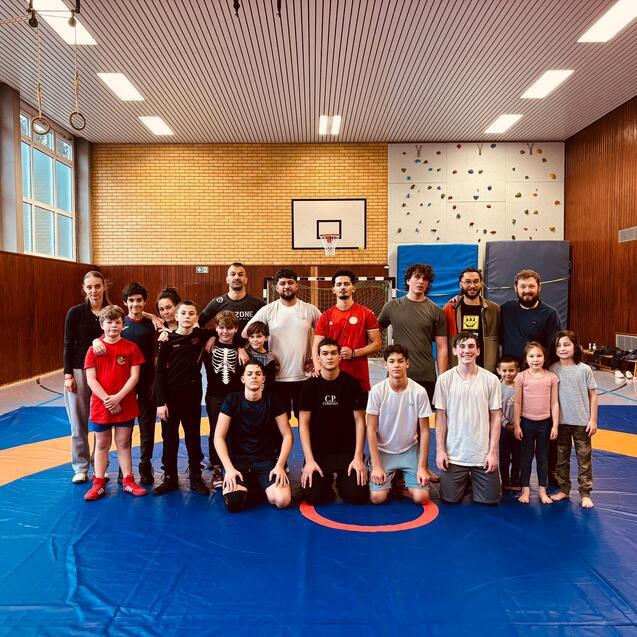 Gruppenfoto von Jugendlichen und Trainern auf einer Mattenfl&auml;che in einer Turnhalle, im Hintergrund sind W&auml;nde und Fenster sichtbar.