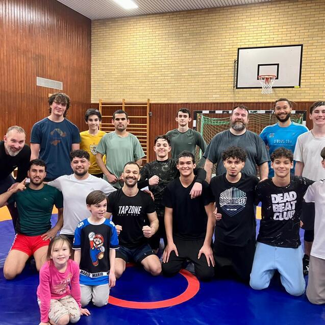 Gruppensfoto von 16 Personen, die auf einer Sportmatte in einem Jugendzentrum stehen, mit l&auml;chelnden Gesichtern und verschiedenen Sportklamotten.