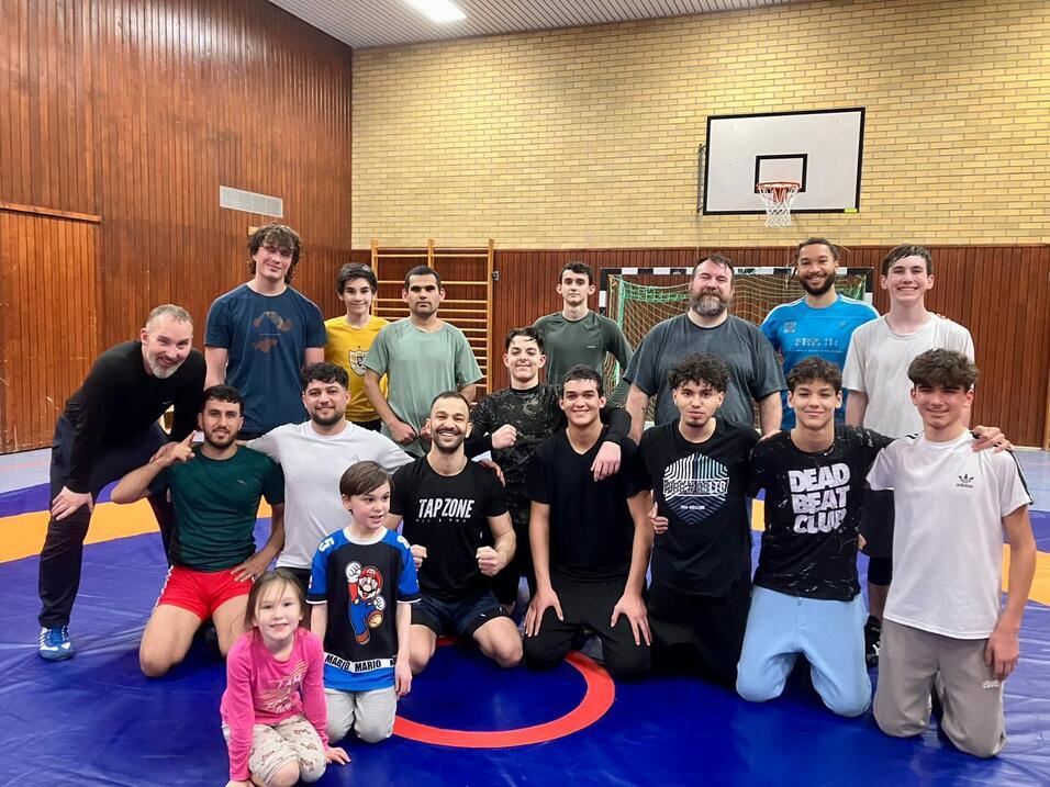 Gruppe von Wrestlern posiert auf einer Matte in einer Sporthalle, darunter ein M&auml;dchen und diverse Erwachsene und Jugendliche.