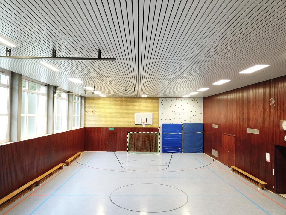 Innenansicht einer Sporthalle mit Basketballkorb, Kletterwand und zwei blauen Matten an der Wand. Holzverkleidung und Fenster.