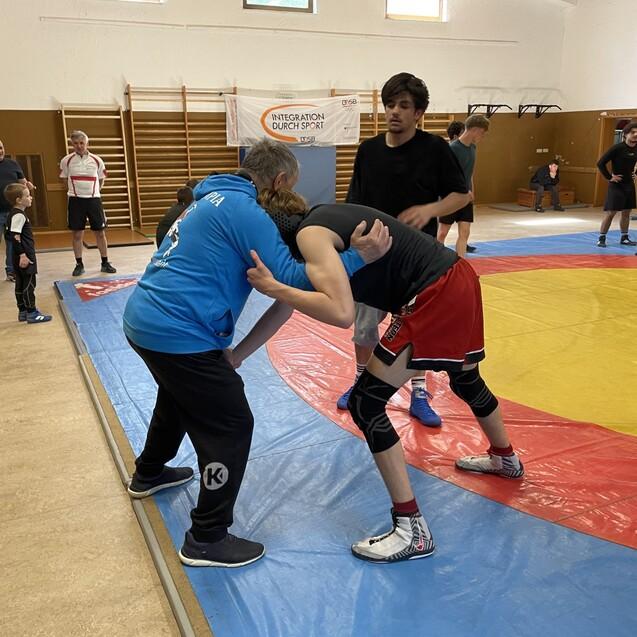 Wrestler im Training auf einer roten Matte, w&auml;hrend ein Trainer Anweisungen gibt. Zuschauer beobachten die &Uuml;bungen.