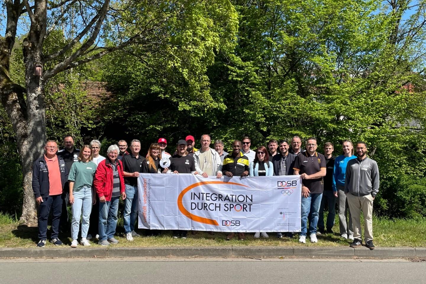 Gruppensch&auml;ffungsfoto mit Banner &bdquo;Integration durch Sport&ldquo; und vielen Teilnehmern vor gr&uuml;ner Kulisse.