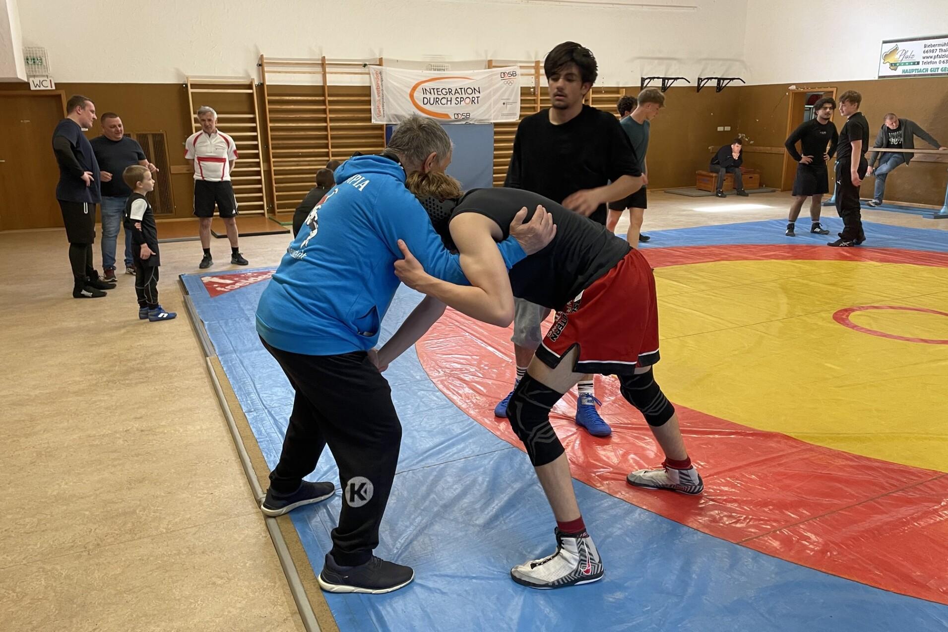 Wrestler im Training auf einer roten Matte, w&auml;hrend ein Trainer Anweisungen gibt. Zuschauer beobachten die &Uuml;bungen.