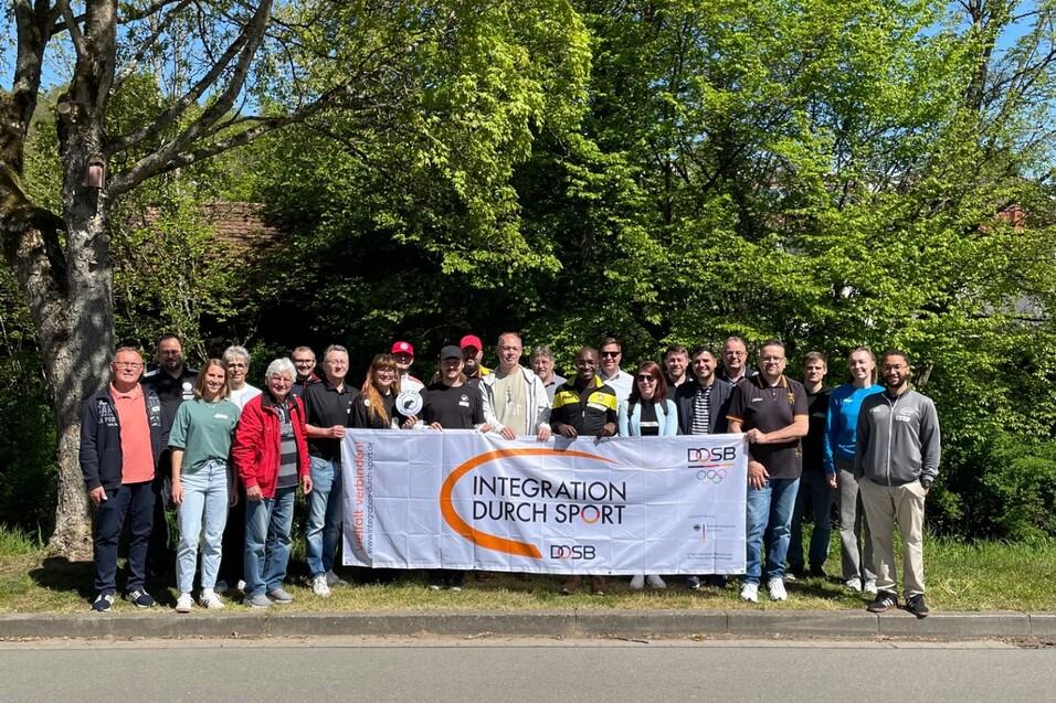 Gruppensch&auml;ffungsfoto mit Banner &bdquo;Integration durch Sport&ldquo; und vielen Teilnehmern vor gr&uuml;ner Kulisse.