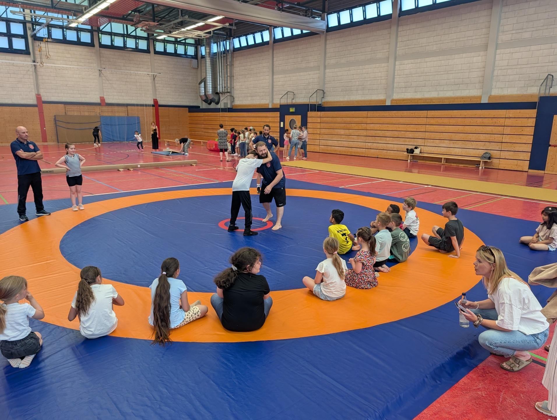 Zwei Kinder &uuml;ben Ringen auf einer Matte, umgeben von beobachtenden Kindern und Trainern in einer Sporthalle.