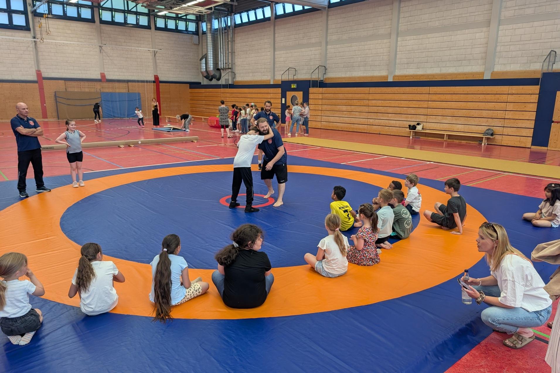 Zwei Kinder &uuml;ben Ringen auf einer Matte, umgeben von beobachtenden Kindern und Trainern in einer Sporthalle.